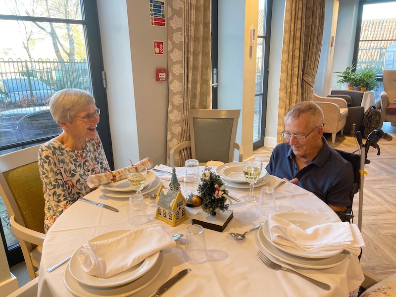 elderly female with leaves on her top and an elderly male with a blue shirt on sat around a dining table for christmas dinner with wine