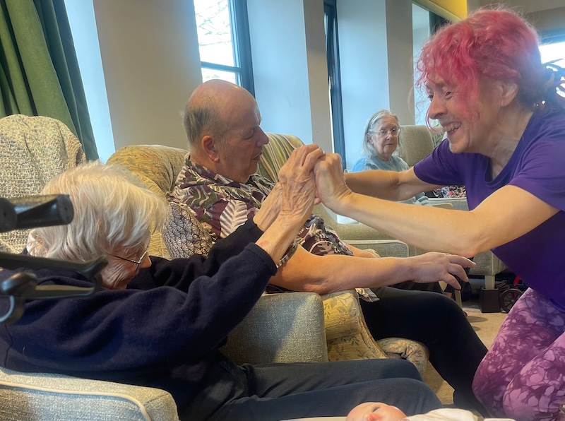 an elderly lady and carer sat holding hands and smiling at each other