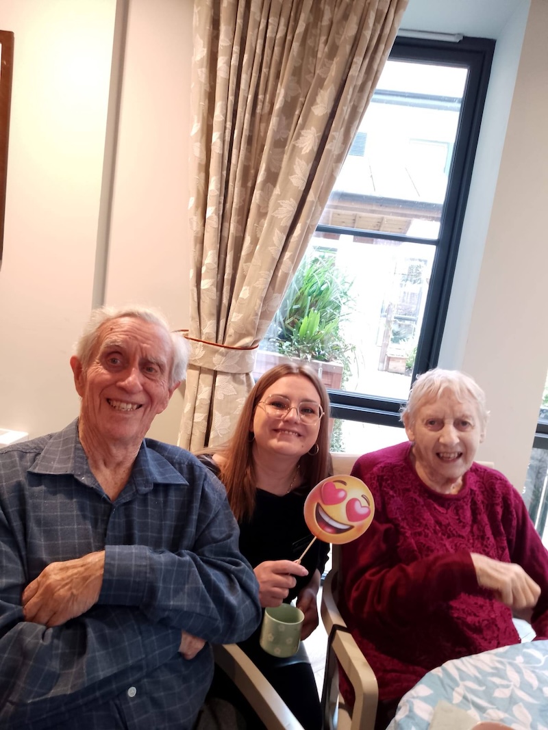 an elderly man and woman sat down next to each other in chairs smiling with a carer between them