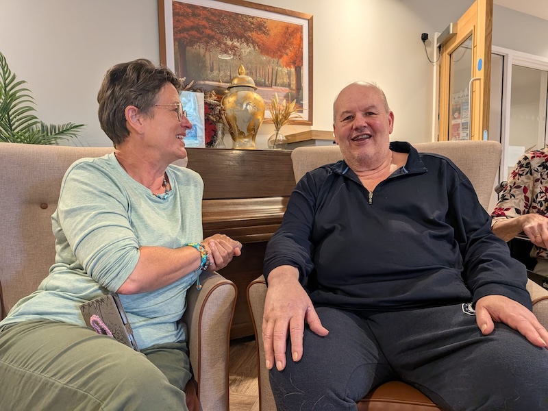 a bald man with a blue jumper on sat in an armchair next to a woman who is looking at him and smiling
